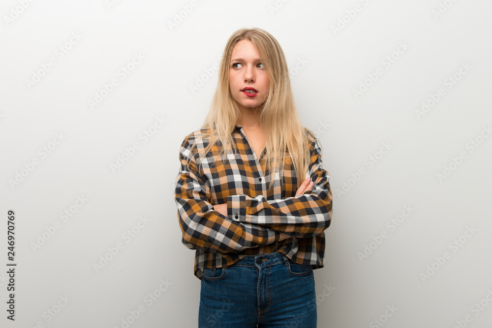 Blonde young girl over white wall with confuse face expression while bites lip Blonde young girl over white wall with confuse face expression while bites lip