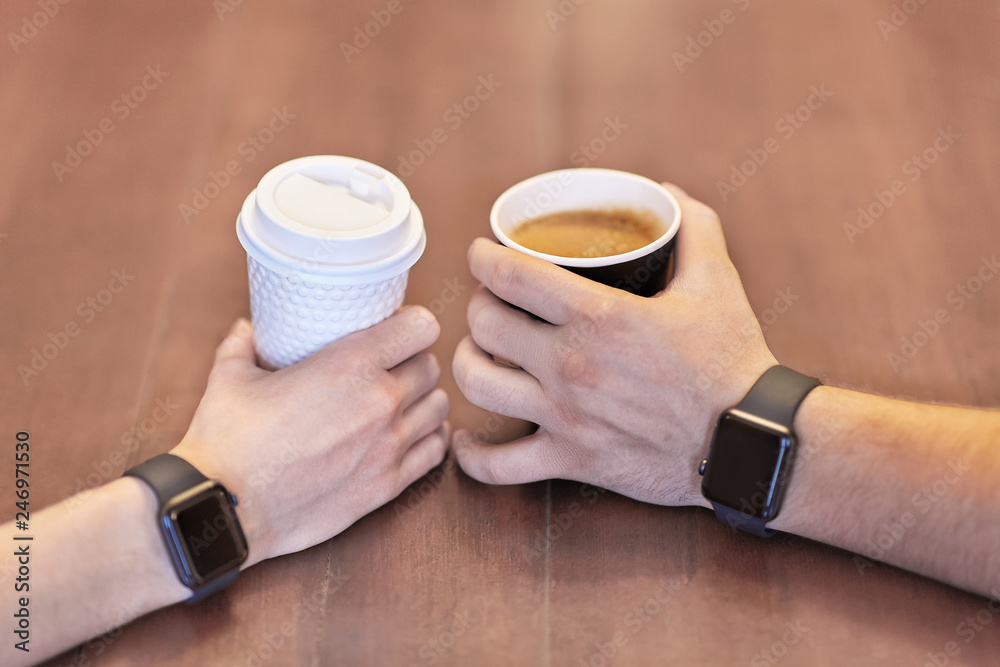 Two hands, male and female, both with equal electronic wrist watches, holding cups of coffee, white and black, on the wooden table. Date or friends meeting. Indoors, copy space.