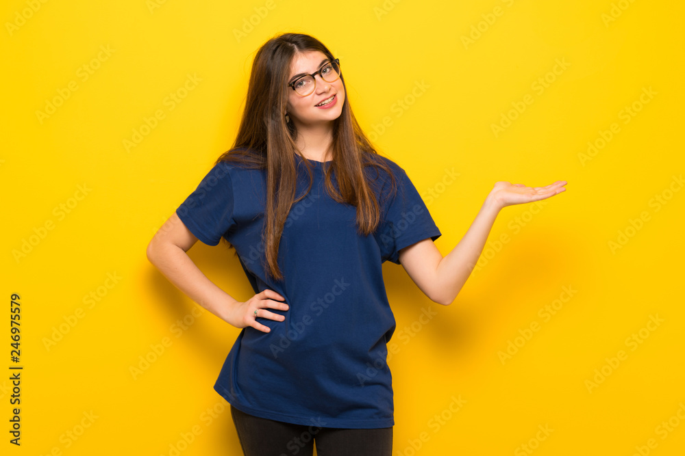 Young woman with glasses over yellow wall holding copyspace imaginary on the palm to insert an ad Young woman with glasses over yellow wall holding copyspace imaginary on the palm to insert an ad