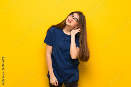Young woman with glasses over yellow wall listening to something by putting hand on the ear © luismolinero
