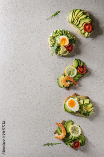 Variety of vegetarian sandwiches with sliced avocado, sun dried tomatoes, egg, shrimps, arugula served over white grey spotted background. Flat lay, space