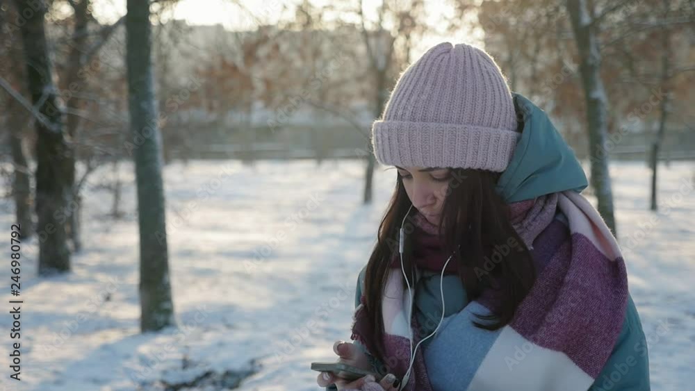 beautiful young woman puts on earphones on the street in the winter park at sunset beautiful young woman puts on earphones on the street in the winter park at sunset