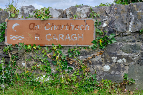 Sign on low road wall for River Caragh, both in English and in Gaelic.  Vine growing on wall and sign photo