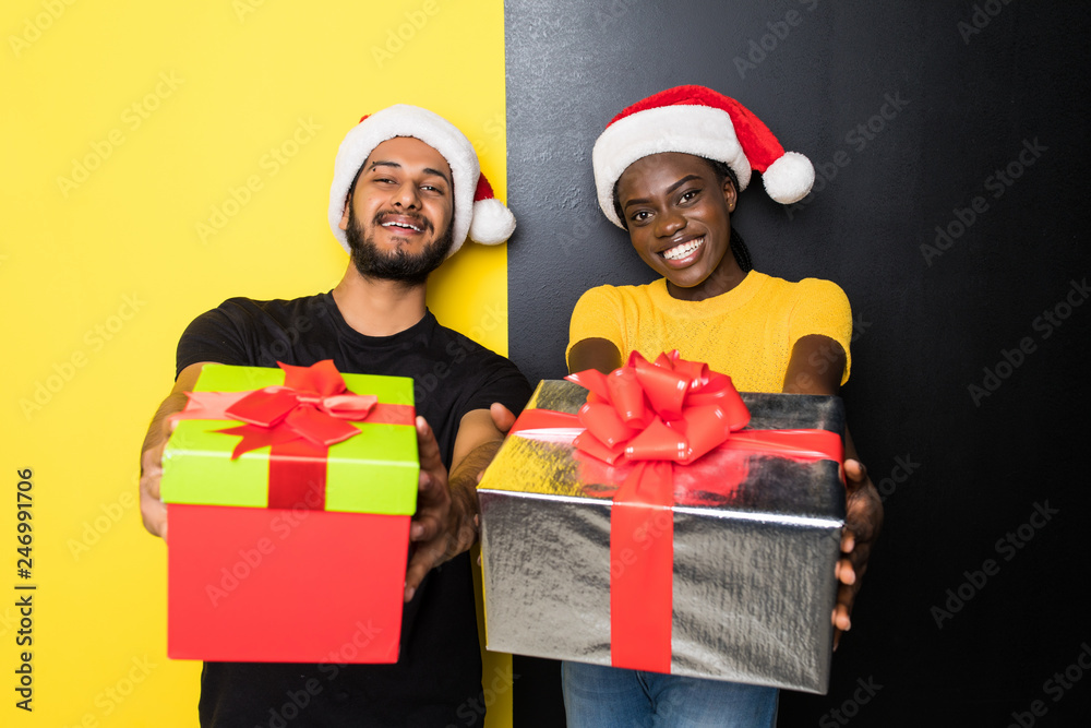 Fototapeta premium Young mixed race couple, indian man and african woman wearing santa hats with Christmas gift standing isolated on yellow black background