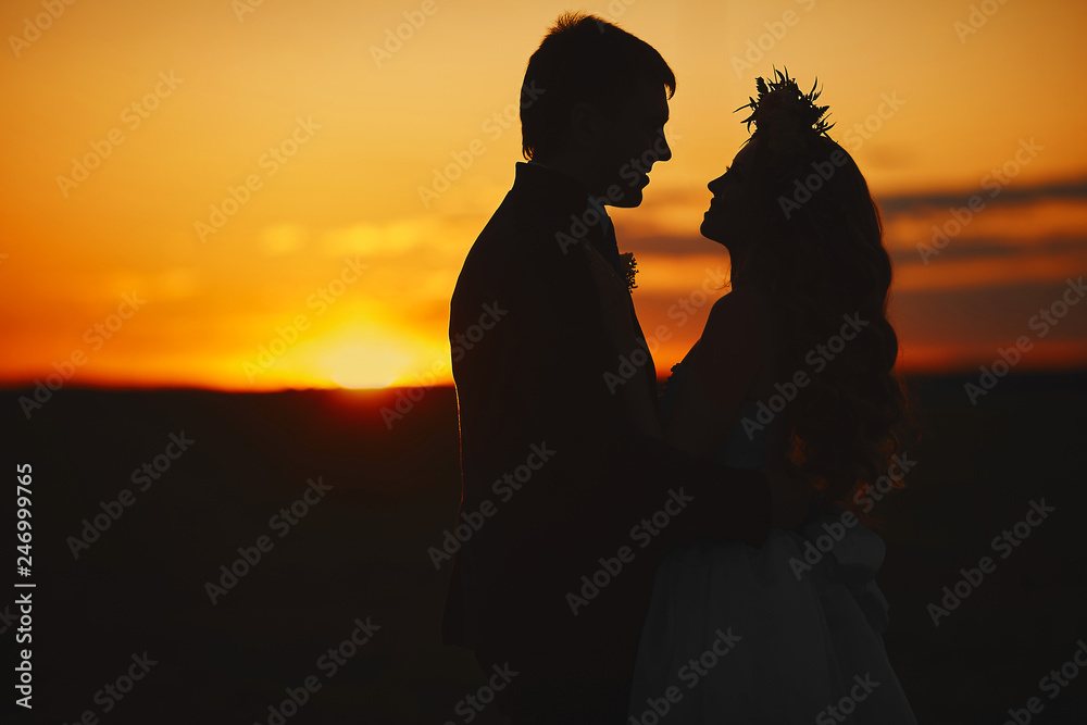 Silhouette of a couple of lovers at the sunset, beautiful blonde model girl with curly hair and with a floral wreath on her head and the young man in stylish suit posing at the sunset