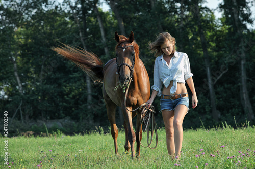 Beautiful cowgirl leading her horse in woods glade at sunset 