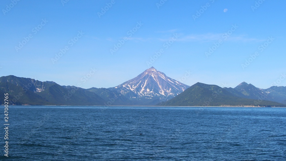 View of Vilyuchinsky volcano (also called Vilyuchik) from water. It's a stratovolcano in the southern part of Kamchatka Peninsula, Russia. Moon is visible in the sky. View of Vilyuchinsky volcano (also called Vilyuchik) from water. It's a stratovolcano in the southern part of Kamchatka Peninsula, Russia. Moon is visible in the sky.