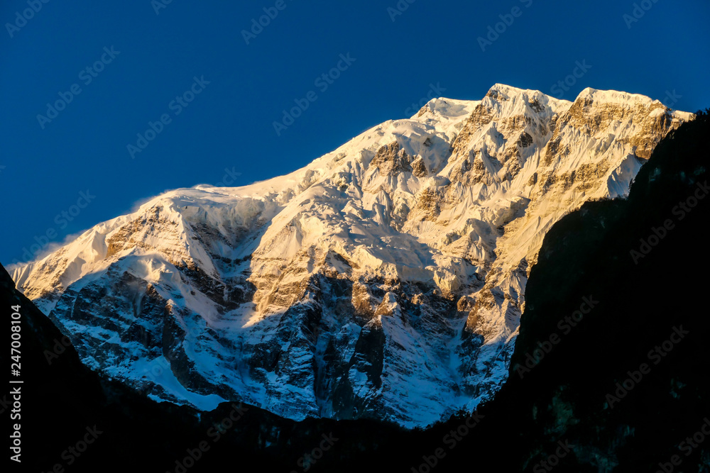 Fototapeta premium Annapurna III captured with the first sun rays on it's peak, during the sunrise. Seen from Chame, Annapurna Circuit Trek, Nepal. Clear sky above the peak. Valley still covered with shadows.