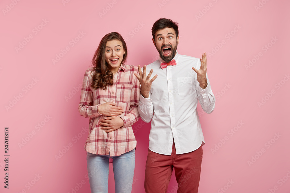 Good news concept. Overjoyed Caucasian man in white shirt and red trousers gestures happily, finds out about wife pregnancy, ready to become father. Positive expectant mother keeps hands on belly Good news concept. Overjoyed Caucasian man in white shirt and red trousers gestures happily, finds out about wife pregnancy, ready to become father. Positive expectant mother keeps hands on belly