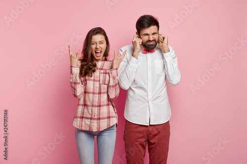 Joyful Caucasian woman makes horn gesture, exclaims as hears loud heavy metal, displeased bearded man plugs ears, doesnt like such music, have different tastes, isolated over pink background.