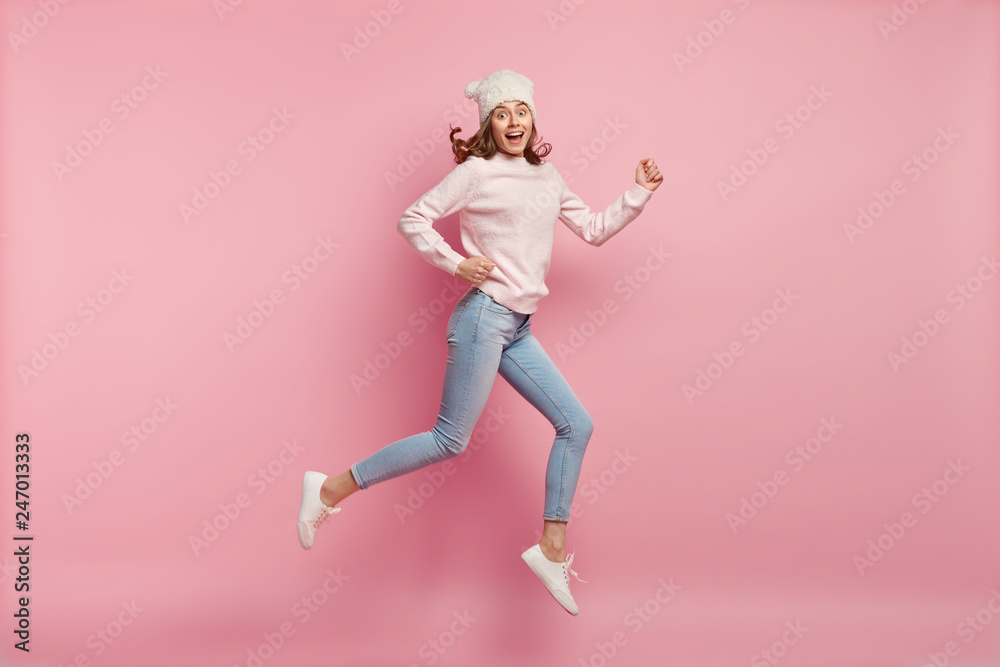 Horizontal shot of happy young Caucasian woman being photographed in motion, rejoices success, expresses positiveness, wears hat, jumper, jeans and sportshoes, jump high against pink background