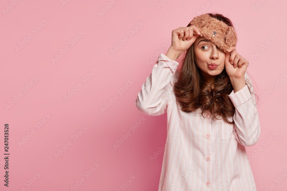 Indoor shot of funny young European woman wears eyemask, pyjamas, looks away, poses against pink background with copy space for your promotional content or advertisement. People and rest concept - obrazy, fototapety, plakaty 