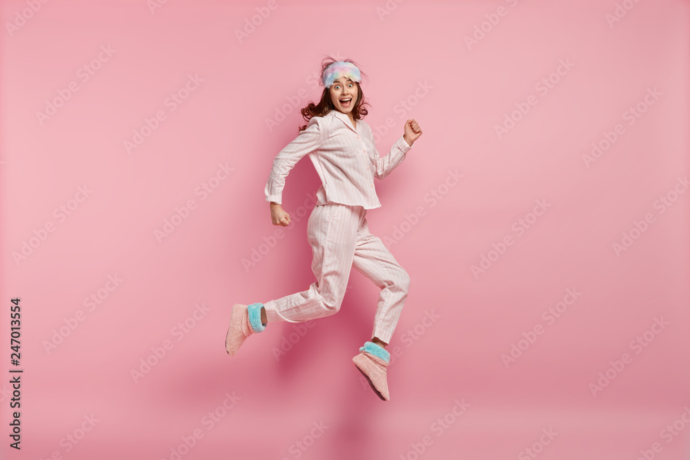 Successful joyful female feels like winner, jumps high in air, wears casual pyjamas and eyemask, wakes up in high spirit, isolated over pink background. People, rest, emotions and sleeping concept - obrazy, fototapety, plakaty 