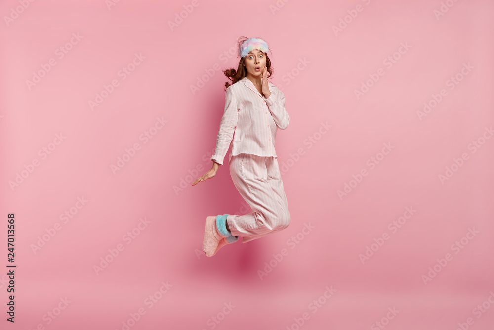 Portrait of surprised young lady wears casual pyjamas, eyemask and warm home boots, jumps in air, shocked to oversleep in morning, isolated over pink background. People and bed time concept.