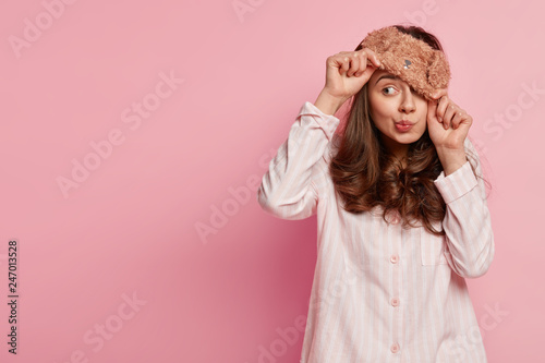 Indoor shot of funny young European woman wears eyemask, pyjamas, looks away, poses against pink background with copy space for your promotional content or advertisement. People and rest concept photo
