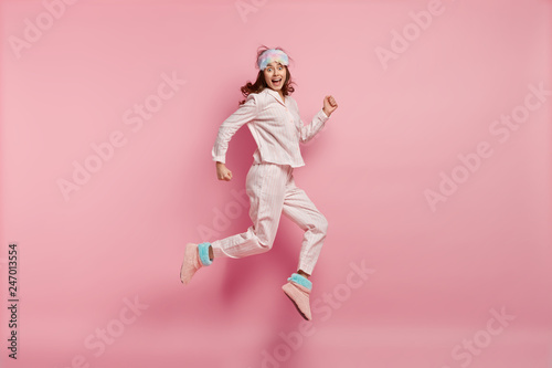 Successful joyful female feels like winner, jumps high in air, wears casual pyjamas and eyemask, wakes up in high spirit, isolated over pink background. People, rest, emotions and sleeping concept