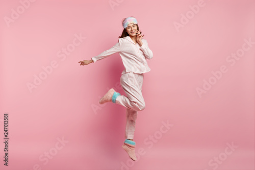 Indoor shot of overjoyed European woman raises leg, spreads hand, wears casual pyjamas and eyemask, smiles happily, isolated over pink background, enjoys bed time and good rest during weekend