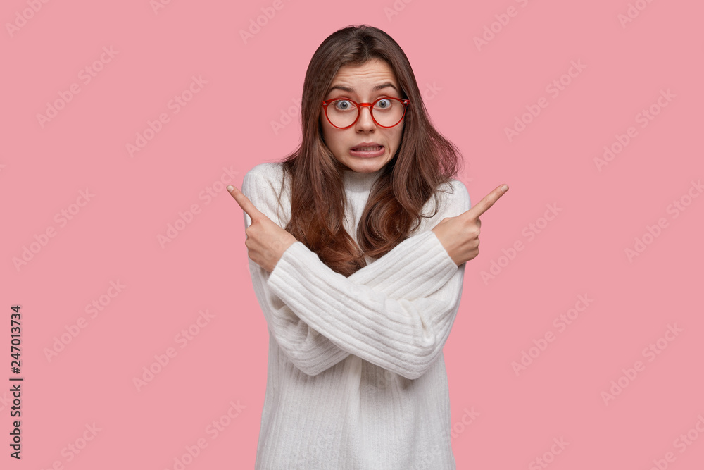 Puzzled European woman crosses hands over chest, clenches teeth, shows direction at different sides, dressed in white clothes, isolated over pink background. Advertisement or promotion concept Puzzled European woman crosses hands over chest, clenches teeth, shows direction at different sides, dressed in white clothes, isolated over pink background. Advertisement or promotion concept