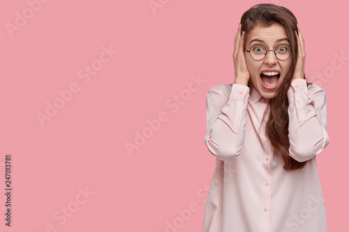Obraz na plátně Emotional young woman in despair keeps hands on ears, opens mouth widely, wears optical spectacles, isolated over pink background with copy space aside for your promotion