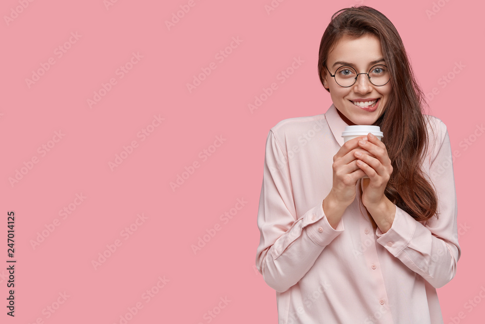 Studio shot of happy young woman with pleased expression, holds disposable cup of coffee, enjoys hot drink and nice weather, isolated over pink kbackground with free space for your advertisement Studio shot of happy young woman with pleased expression, holds disposable cup of coffee, enjoys hot drink and nice weather, isolated over pink kbackground with free space for your advertisement