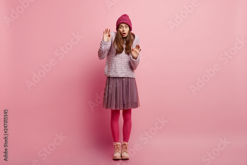 Horizontal shot of surprised frightened young lady keeps hands in protective gesture, afraids of phobia wears warm knitted hat, jumper full skirt, pink pantyhose and boots, models indoor. Fear concept