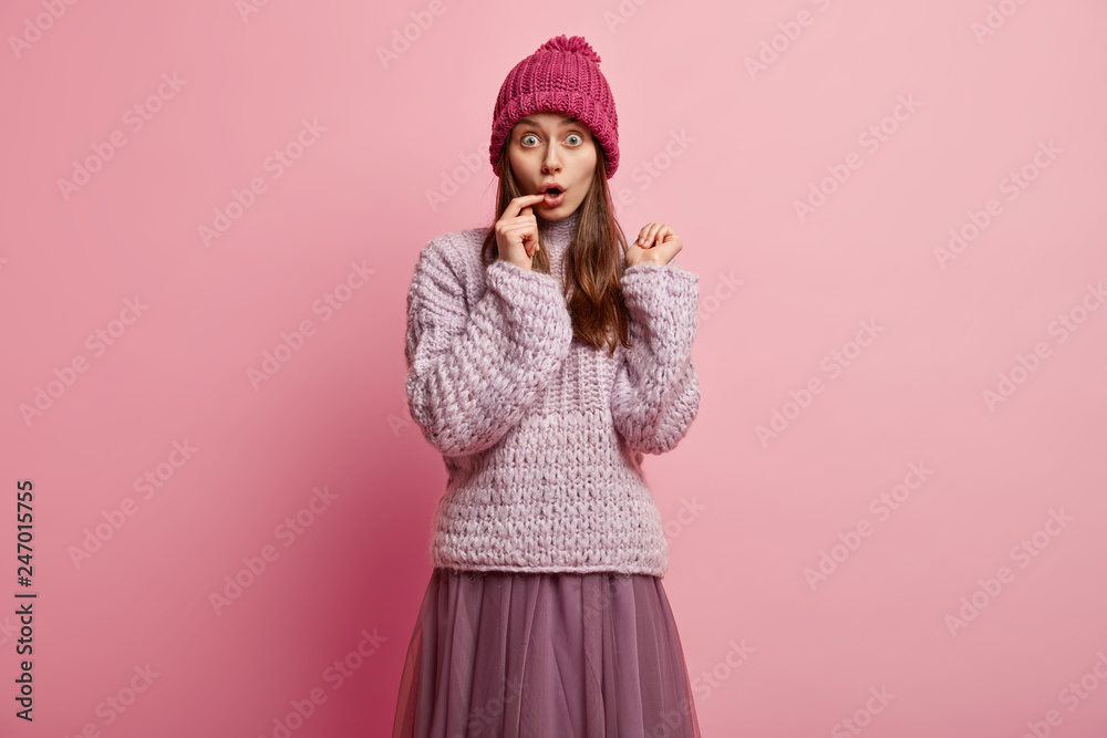 Puzzled scared hipster girl has bugged eyes, keeps fore finger on lips, wears winter hat, oversized long sleeved jumper, skirt, models over rosy background, has overwhelmed facial expression Puzzled scared hipster girl has bugged eyes, keeps fore finger on lips, wears winter hat, oversized long sleeved jumper, skirt, models over rosy background, has overwhelmed facial expression