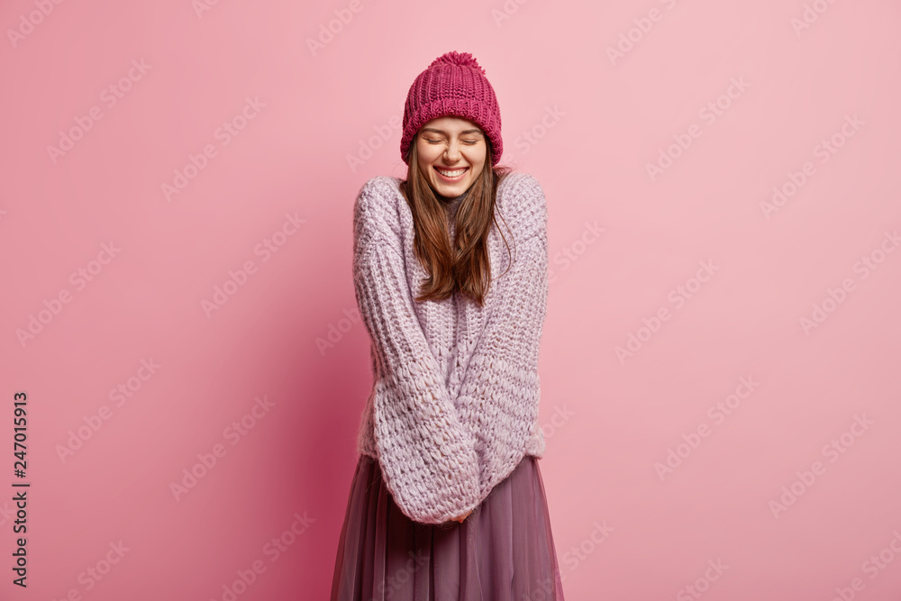 Positive Caucasian young female model keeps hands together, wears long sleeved knitted sweater, pink hat, closes eyes from pleasure, models over rosy background. People and emotions concept. Positive Caucasian young female model keeps hands together, wears long sleeved knitted sweater, pink hat, closes eyes from pleasure, models over rosy background. People and emotions concept.