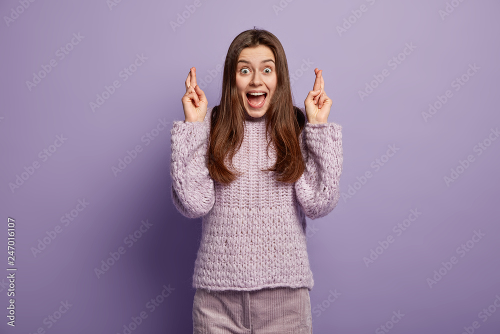 Positive overjoyed woman eagers dreams come true, has fingers crossed, dressed in knitted sweater, opens mouth widely, isolated over purple background, believes in good luck. People and wish Positive overjoyed woman eagers dreams come true, has fingers crossed, dressed in knitted sweater, opens mouth widely, isolated over purple background, believes in good luck. People and wish