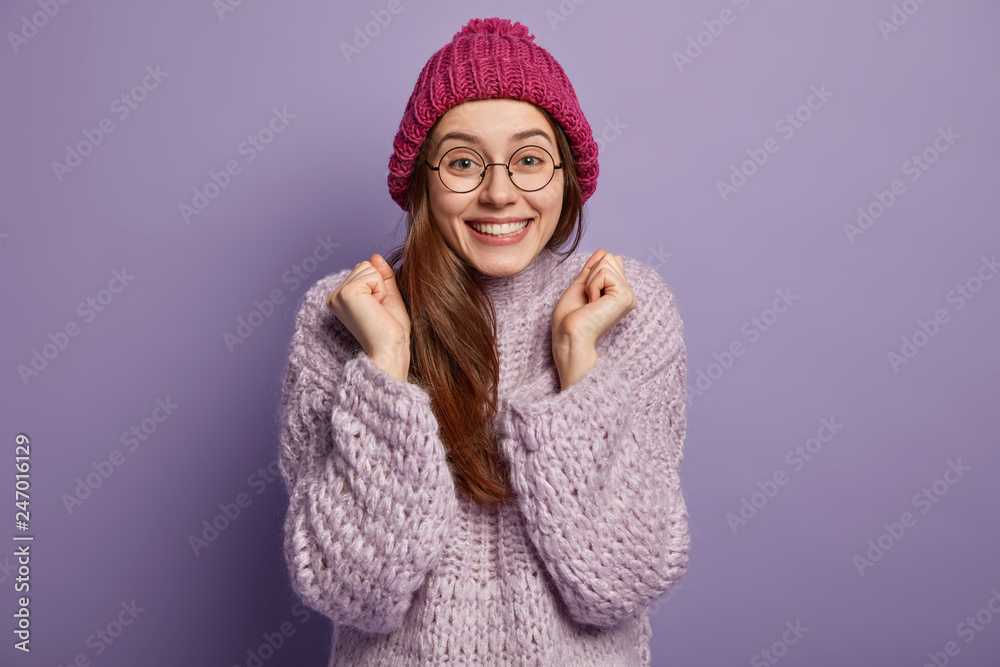 Happy pleased woman clenches fists, smiles broadly, wears bright pink knitted hat and loose warm sweater, rejoices good weather, models over purple background. Pretty girl in winter clothes.