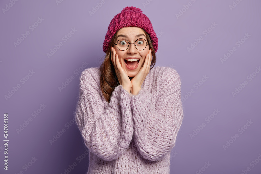 Positive young teenage girl touches both cheeks with palms, stares with widely opened eyes, wears pink knitted hat and oversized sweater, rejoices having fine vacation during winter holidays Positive young teenage girl touches both cheeks with palms, stares with widely opened eyes, wears pink knitted hat and oversized sweater, rejoices having fine vacation during winter holidays