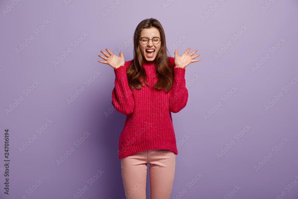 Shot of overjoyed happy young female model spreads palms near face, exclaims with positive emotions, closes eyes from pleasure, looks excited, dressed in red loose sweater, isolated on purple wall Shot of overjoyed happy young female model spreads palms near face, exclaims with positive emotions, closes eyes from pleasure, looks excited, dressed in red loose sweater, isolated on purple wall