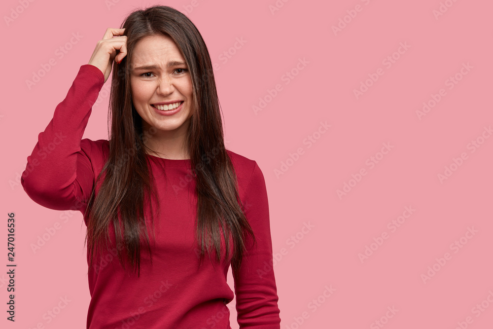 Confused clueless woman cannot understant some hints, scrathes head and looks questioned, wears red jumper, tries to recollect important information in mind, isolated on pink wall with empty space Confused clueless woman cannot understant some hints, scrathes head and looks questioned, wears red jumper, tries to recollect important information in mind, isolated on pink wall with empty space