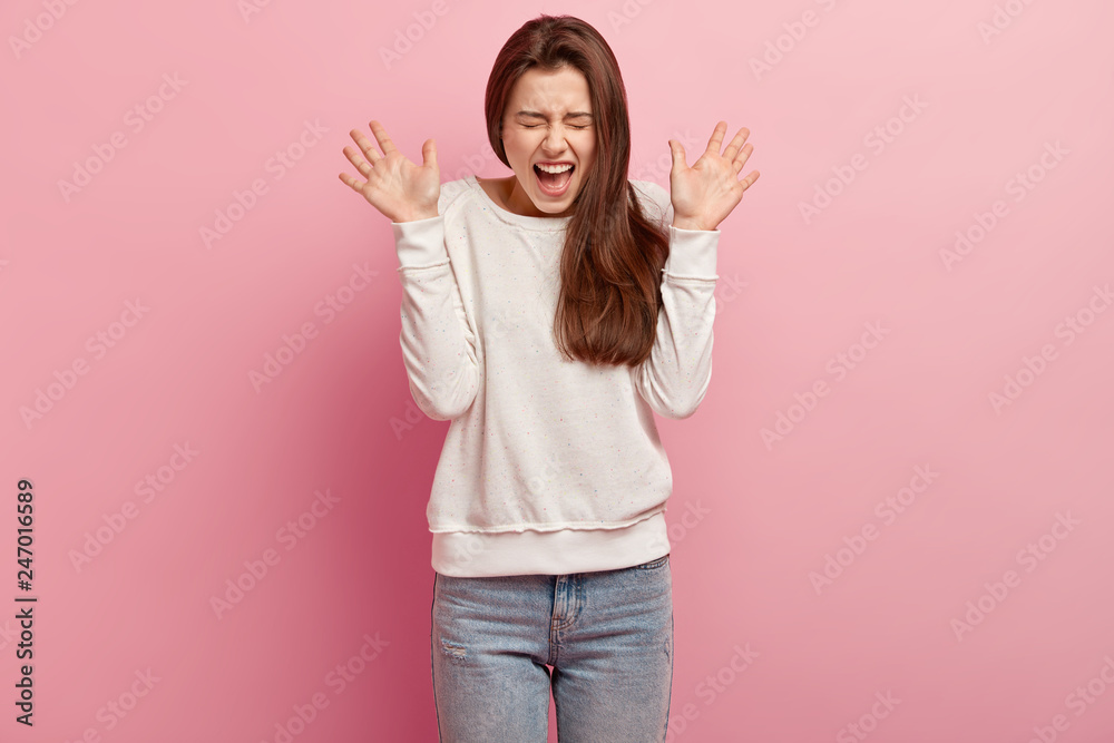 Photo of attractive young woman with dark long hair, raises palms, exclaims happily, feels glad and overemotive, wears casual jumper and jeans isolated over rosy studio background. People, joy concept Photo of attractive young woman with dark long hair, raises palms, exclaims happily, feels glad and overemotive, wears casual jumper and jeans isolated over rosy studio background. People, joy concept