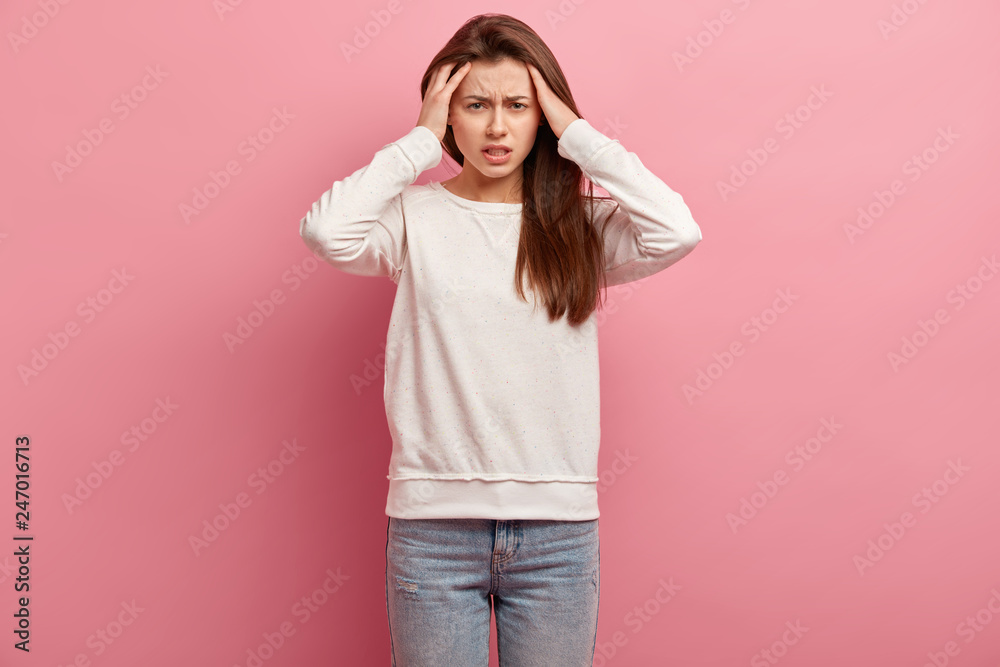 Pretty young European girl keeps hands on head, suffers from migraine, dressed in casual jumper and jeans, stands against pink background, feels under pressure, has headache, disappointment. Pretty young European girl keeps hands on head, suffers from migraine, dressed in casual jumper and jeans, stands against pink background, feels under pressure, has headache, disappointment.