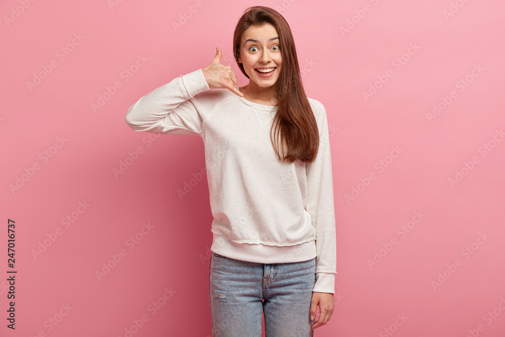 Cheerful woman makes call gesture, dressed in casual pullover, jeans, smiles positively, reminds to be in contact, poses over rosy studio wall. People, communication on distance and body language Cheerful woman makes call gesture, dressed in casual pullover, jeans, smiles positively, reminds to be in contact, poses over rosy studio wall. People, communication on distance and body language