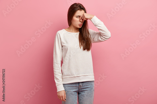 Fotografia Displeased woman covers nose with hand, smells something awful, pinches nose, frowns in displeasure, sees pile of garbage, dressed in casual clothes, isolated over pink background