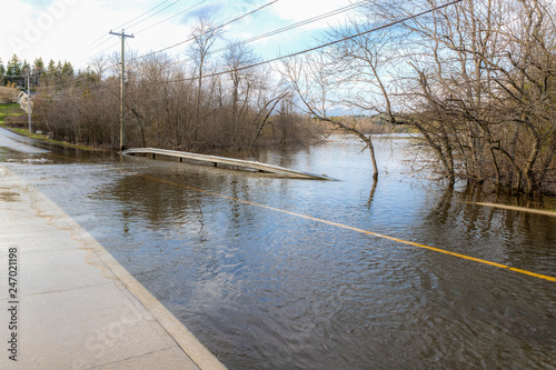 Water covering a flooded road. The yellow dividing line and sidewalk can be seen submerged. Trees in the background.