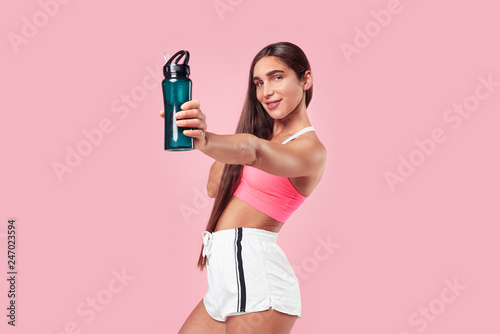 Woman with long hair muscular body in sports clothing relaxing after workout, showing botle of water on pink background. photo