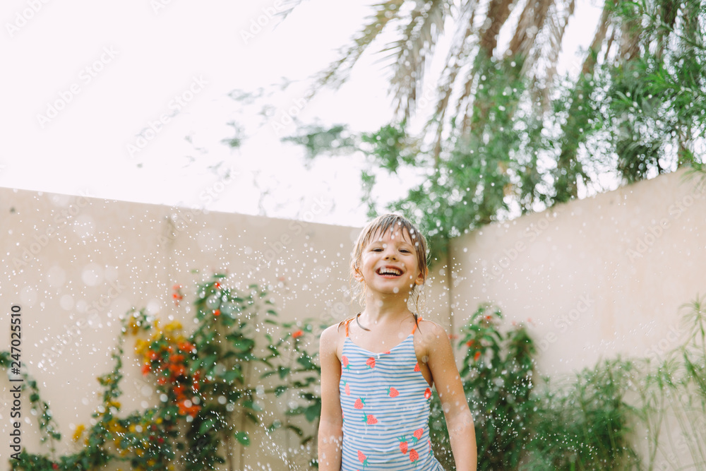 Funny laughing little girl running though garden sprinkler playing with water splashes in the backyard on a sunny hot summer vacation day . Funny laughing little girl running though garden sprinkler playing with water splashes in the backyard on a sunny hot summer vacation day .