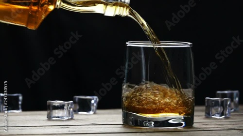 Whiskey pouring into a glass with ice cubes on old wooden table in slow motion. Seamless cinemagraph video