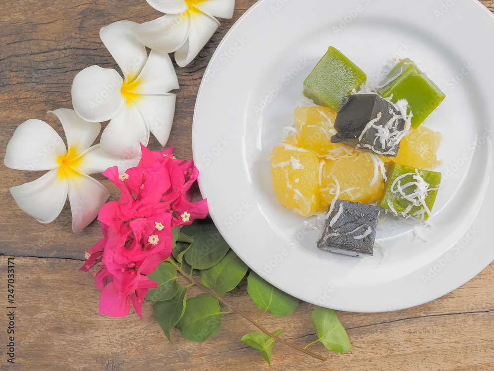 Delicious Thai Dessert Khanom Piek Poon (Burnt Coconut Jelly), Khanom Chun (Thai Jello) and Khanom Man (Coconut Cassava Cake) in white plate with flowers on wood texture background. Delicious Thai Dessert Khanom Piek Poon (Burnt Coconut Jelly), Khanom Chun (Thai Jello) and Khanom Man (Coconut Cassava Cake) in white plate with flowers on wood texture background.