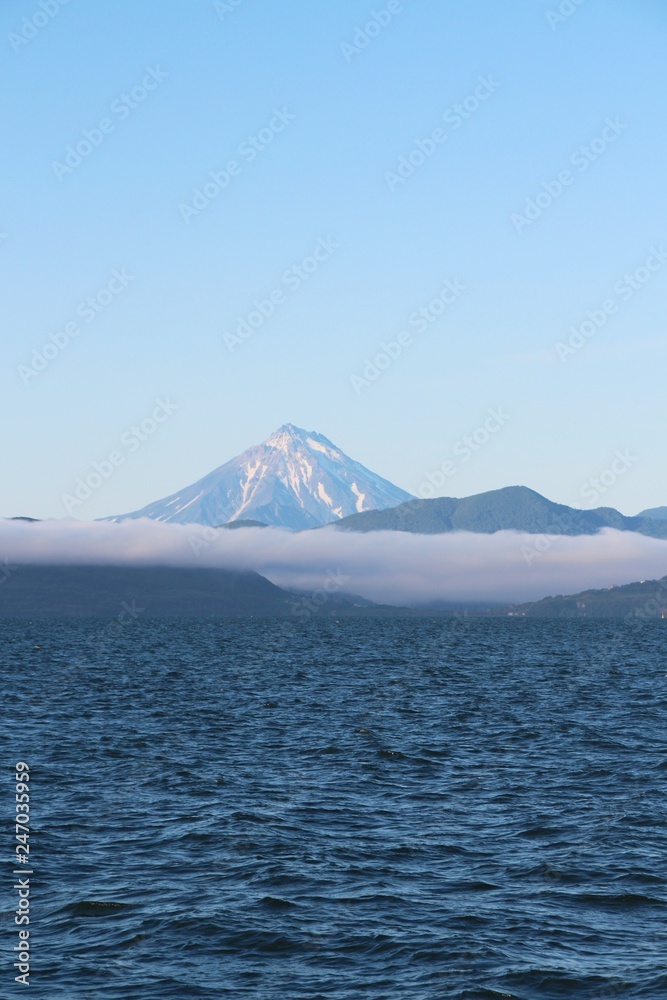 View of Vilyuchinsky volcano (also called Vilyuchik) from tourist boat. The cloud lies on the coastal cliffs. Vilyuchinsky is a stratovolcano in the southern part of Kamchatka Peninsula, Russia. View of Vilyuchinsky volcano (also called Vilyuchik) from tourist boat. The cloud lies on the coastal cliffs. Vilyuchinsky is a stratovolcano in the southern part of Kamchatka Peninsula, Russia.