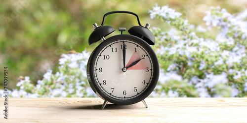 European daylight saving time. Alarm clock on wooden desk, blur spring nature background. 3d illustration