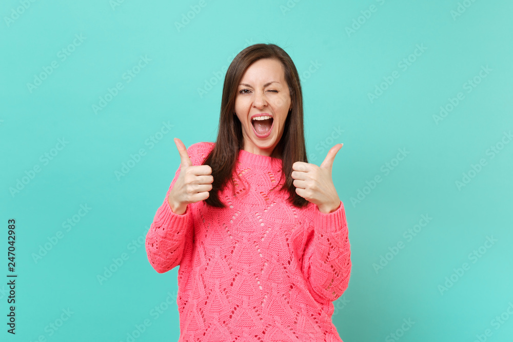 Funny young woman in knitted pink sweater blinking and showing thumbs up isolated on blue turquoise wall background, studio portrait. People sincere emotions, lifestyle concept. Mock up copy space. Funny young woman in knitted pink sweater blinking and showing thumbs up isolated on blue turquoise wall background, studio portrait. People sincere emotions, lifestyle concept. Mock up copy space.