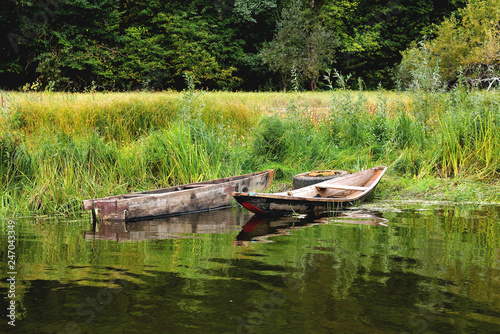 Two old boats on the shore are tied to an old car wheel, on a background of green grass a sunny summer day