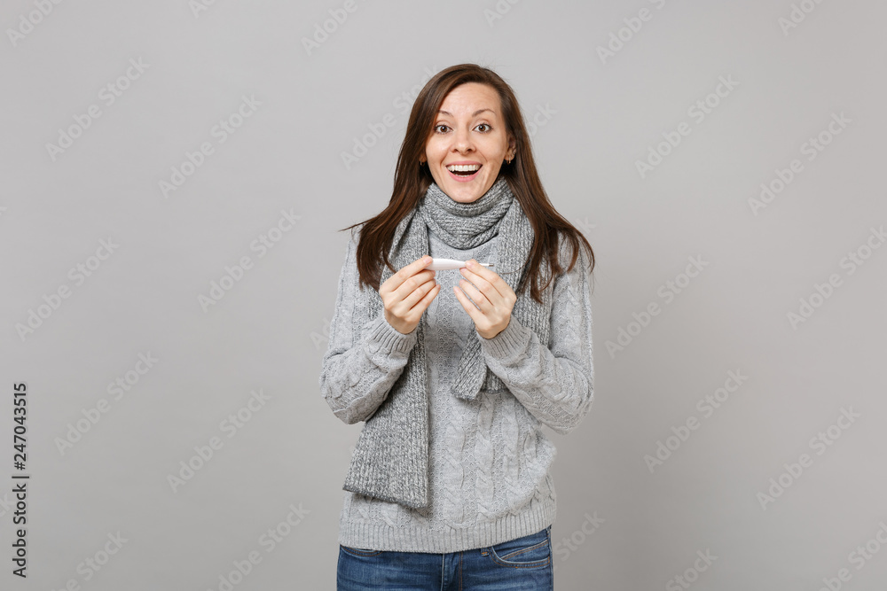 Joyful excited young woman in gray sweater scarf holding thermometer isolated on grey wall background in studio. Healthy lifestyle, ill sick disease treatment, cold season concept. Mock up copy space.