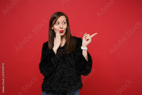 Confused young woman in black fur sweater pointing index finger aside putting hand on cheek isolated on bright red background in studio. People sincere emotions, lifestyle concept. Mock up copy space.