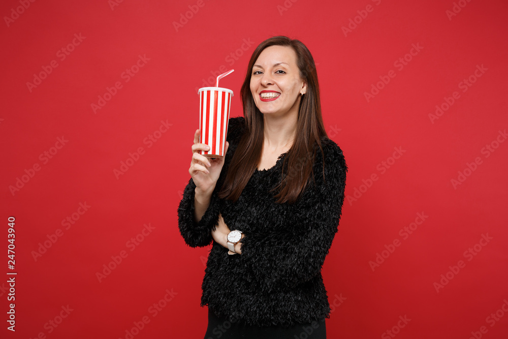 Portrait of smiling young girl in black fur sweater holding plastic cup of cola or soda isolated on bright red wall background in studio. People sincere emotions lifestyle concept. Mock up copy space. - obrazy, fototapety, plakaty 