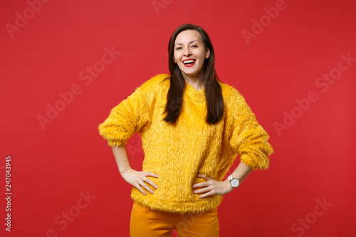 Portrait of laughing pretty young woman in yellow fur sweater standing with arms akimbo isolated on bright red wall background in studio. People sincere emotions lifestyle concept. Mock up copy space.
