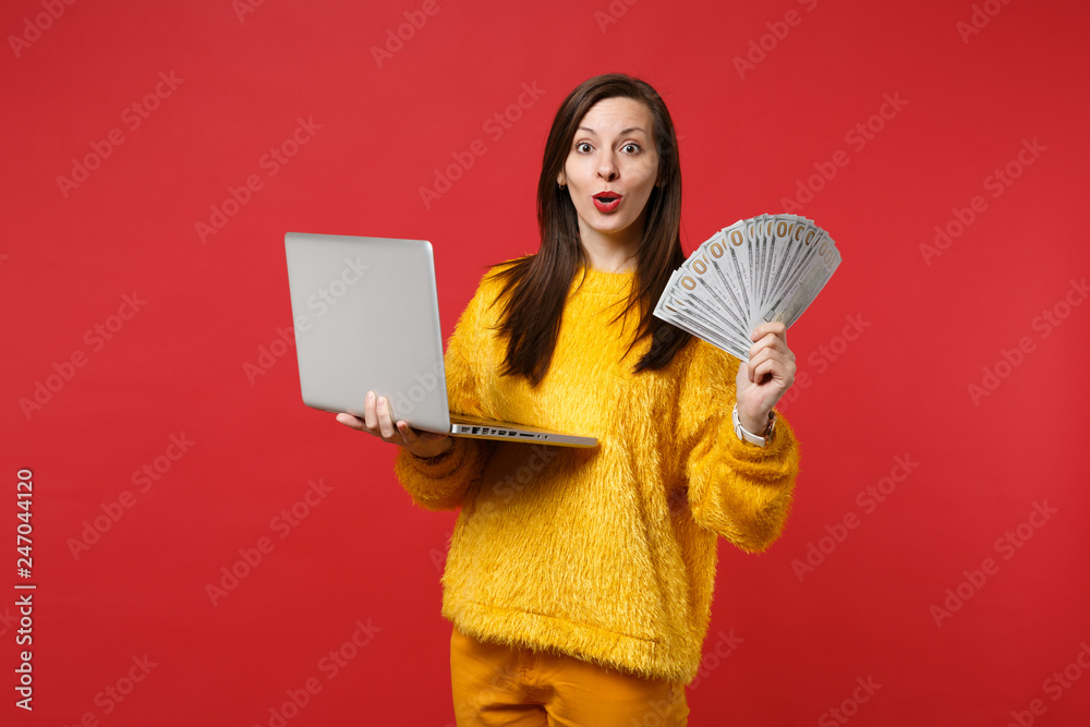 Amazed young woman in yellow fur sweater holding laptop pc computer, fan of money in dollar banknotes, cash money isolated on red background. People emotions, lifestyle concept. Mock up copy space. Amazed young woman in yellow fur sweater holding laptop pc computer, fan of money in dollar banknotes, cash money isolated on red background. People emotions, lifestyle concept. Mock up copy space.